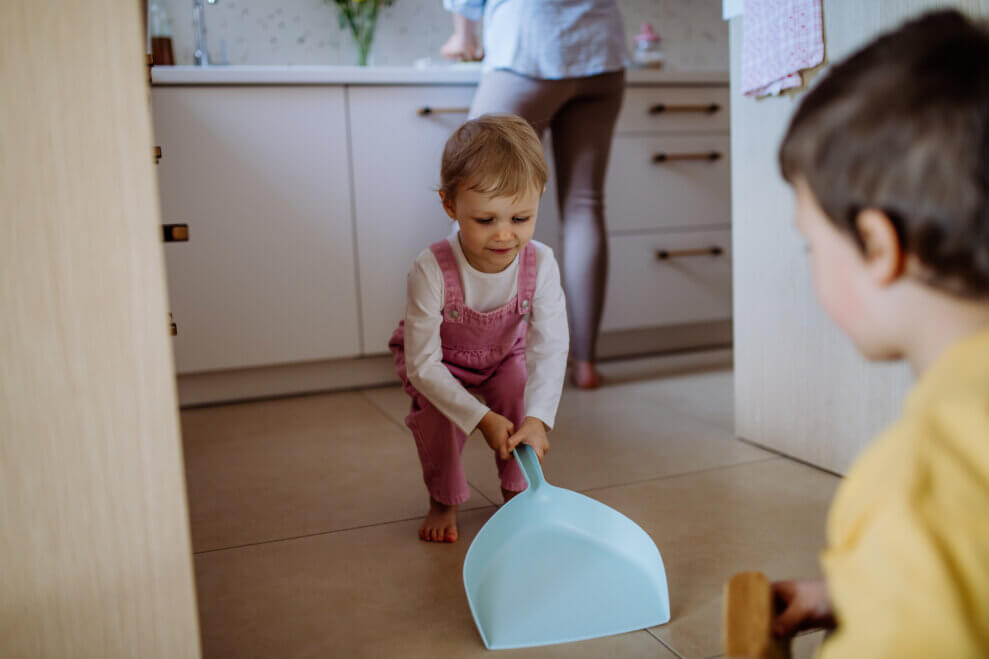 A toddler holds a dustpan against the kitchen floor while her older brother sweeps dirt into it. 
