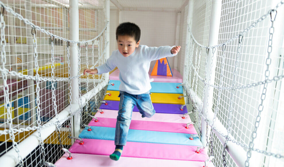 A boy runs across a padded bridge at an indoor playground.