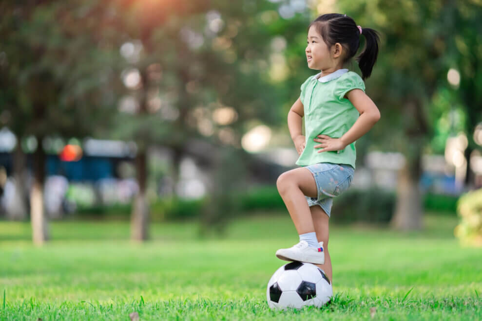 A young girl stands in a park with her foot on a soccer ball and her hands on her hips. She's smiling.