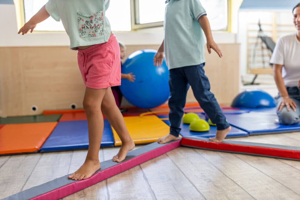 Two children walk along a low foam balance beam with their arms out for balance.