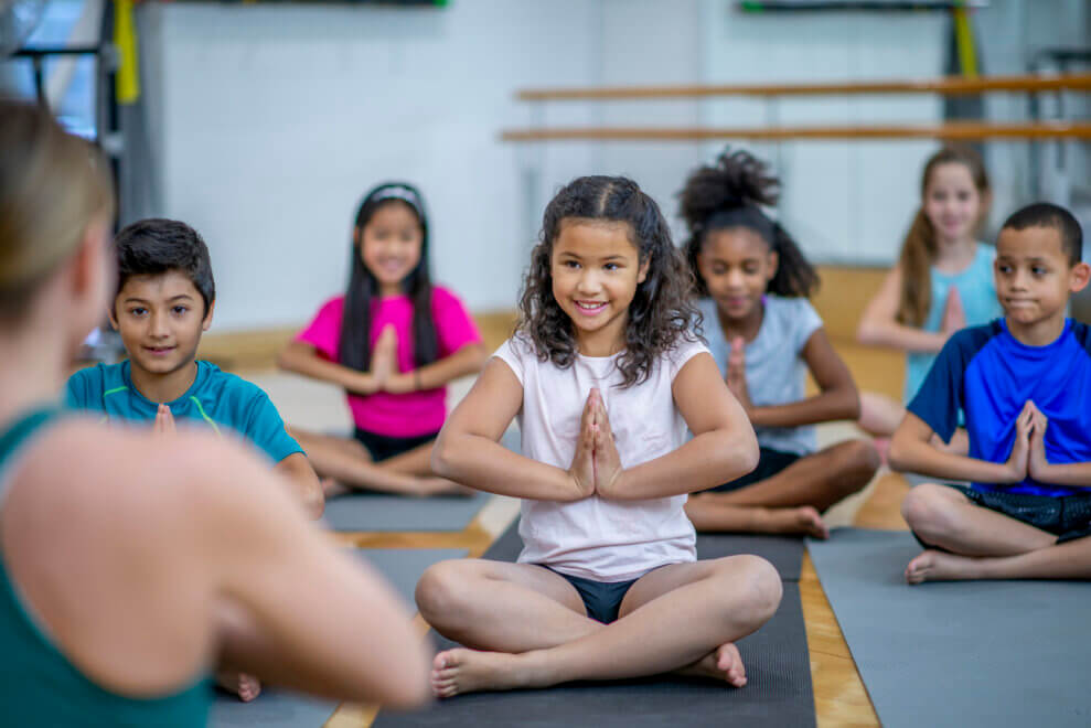 A yoga teacher leads a group of kids in an indoor yoga class. They sit on their mats and hold their hands at heart centre.