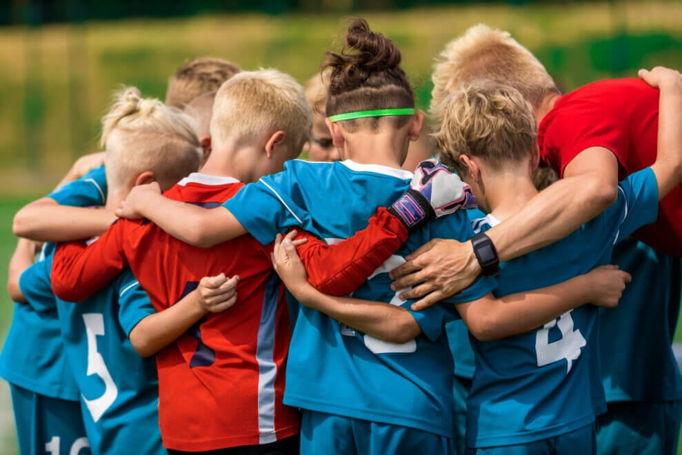 A soccer team of children gathers for a huddle with their arms around each other.