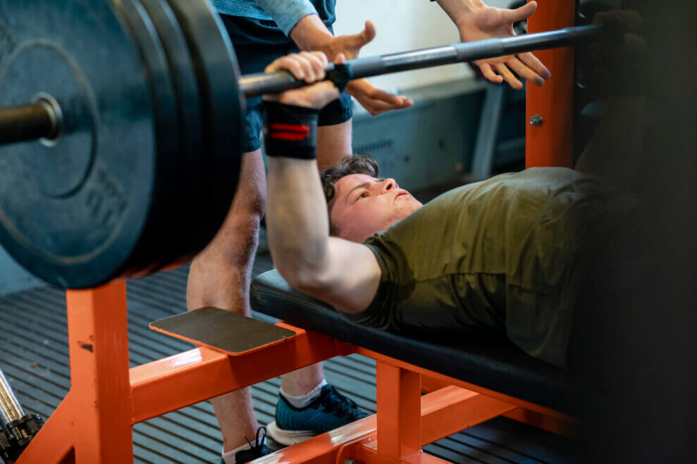 A teenage boy bench presses with a barbell at the gym as an instructor stands over him and spots him. 