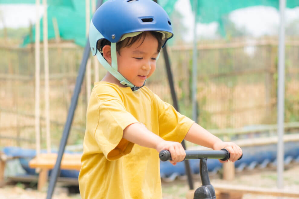 A toddler boy stands over his balance bike with his hands on the handlebars and a smile on his face.