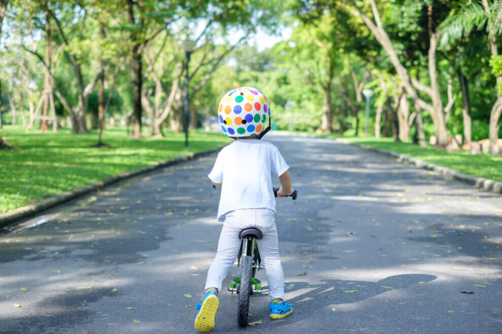 A toddler boy rides his balance bike along a path through a grassy park on a sunny summer day.