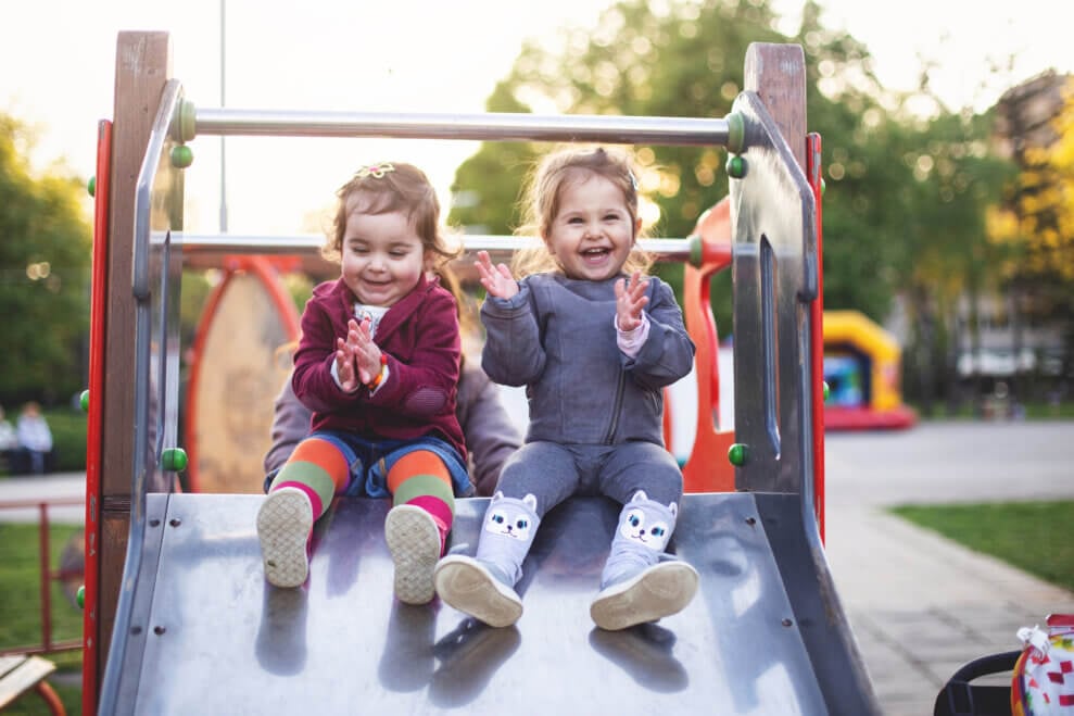 Two toddler girls sit at the top of a playground slide, laughing and clapping their hands.