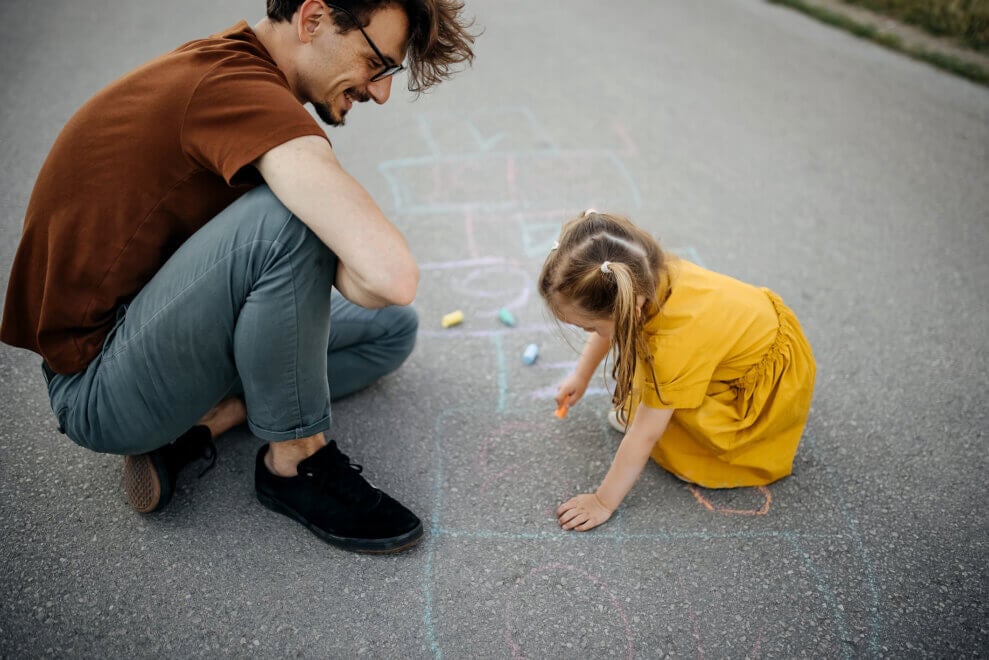 A father and his toddler daughter crouch on the pavement and draw with sidewalk chalk.