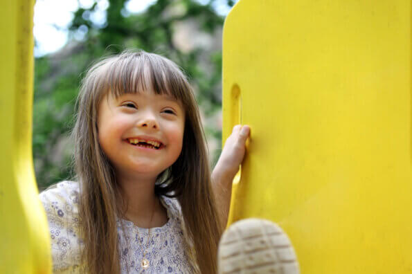 Young girl with Down syndrome sits on a slide at a playground and smiles.