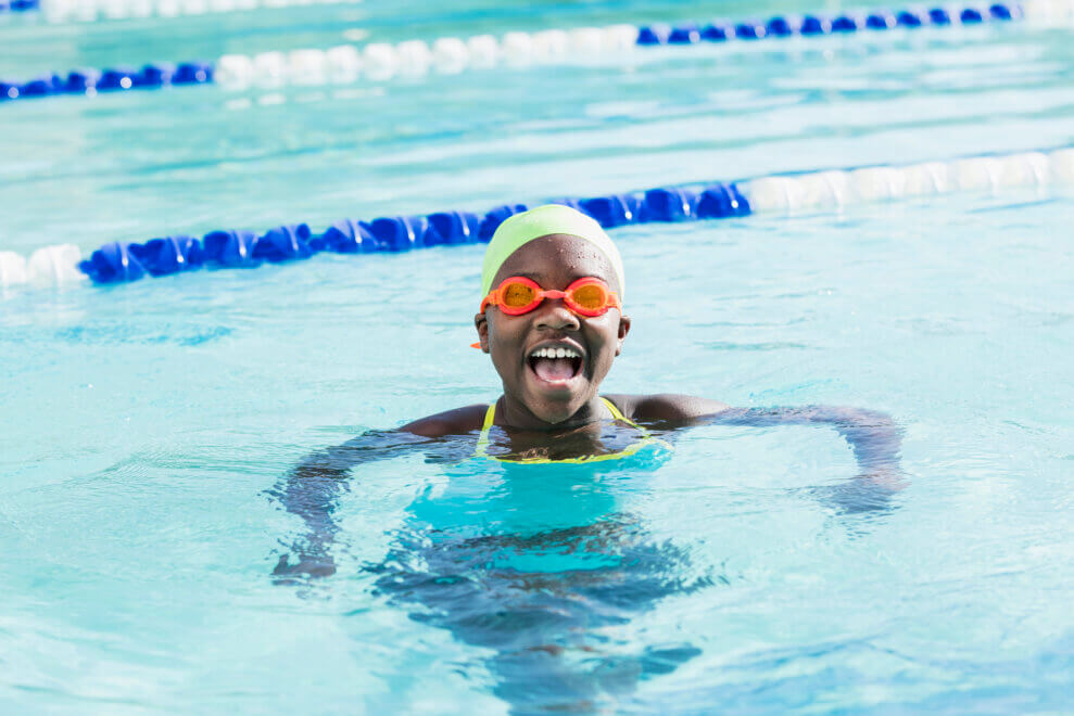 A girl wearing a swim cap and goggles treads water in a swimming pool. She has a big smile on her face.