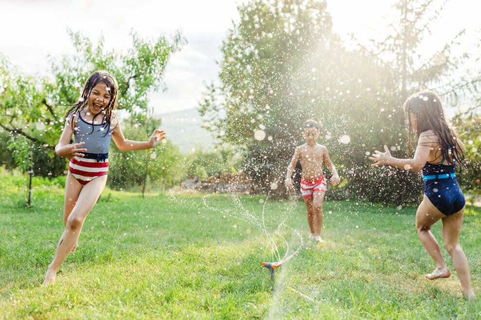 Three children run through a sprinkler on the lawn, laughing and screaming.