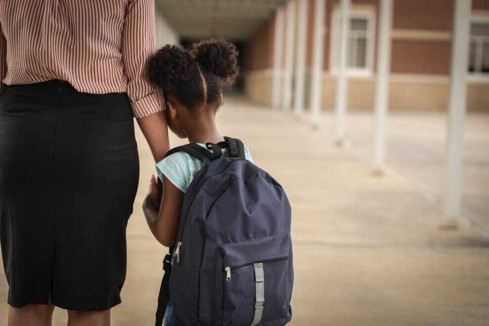 A young girl wearing a backpack holds tight to her mom's hand as they stand in front of a school.
