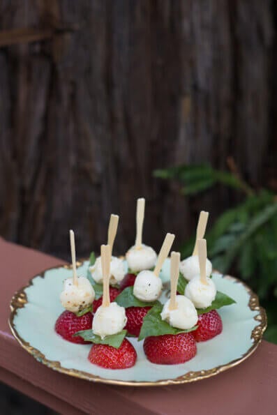 On an outdoor platter, halved strawberries have basil and mozzarella cheese on top of them. 
