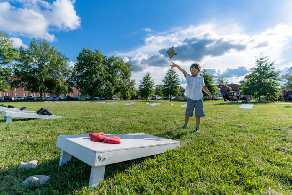 A boy tosses bean bags at a cornhole board on a sunny day in the park.