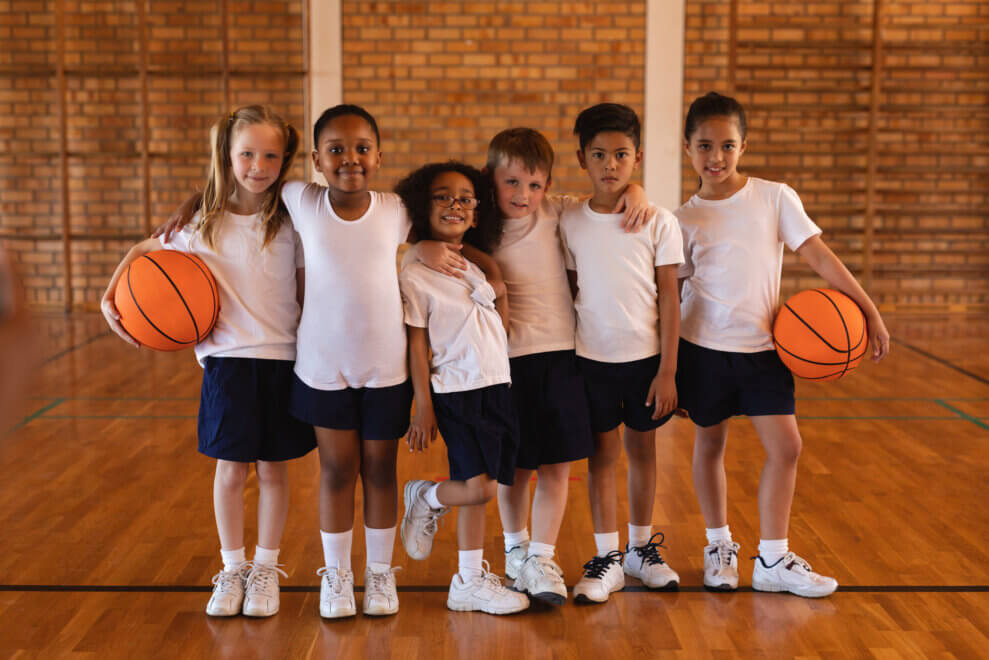 A group of children look at the camera on a basketball court. Two of them hold basketballs.