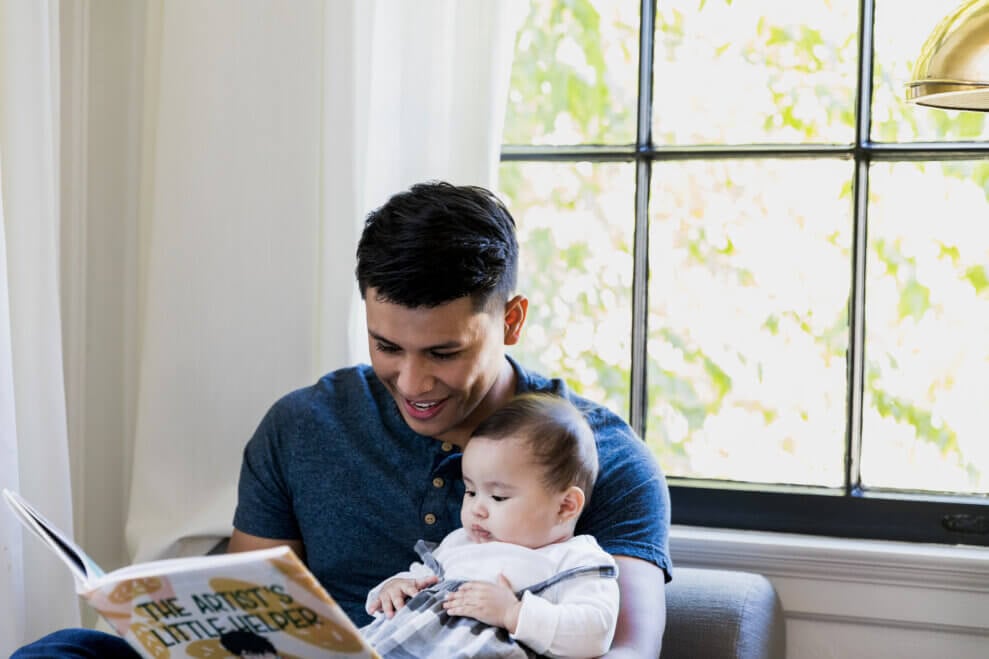 A father reads a book aloud to his baby daughter as they sit in a chair in front of a window in their home.