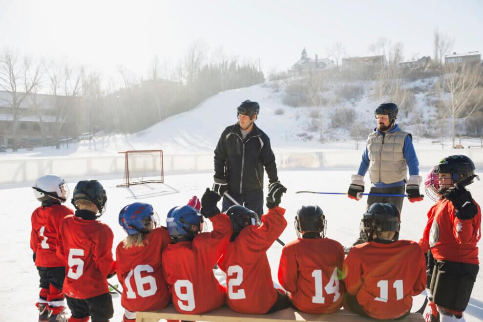 A kids' hockey team sits on the bench next to an outdoor rink as their two coaches give them instructions.