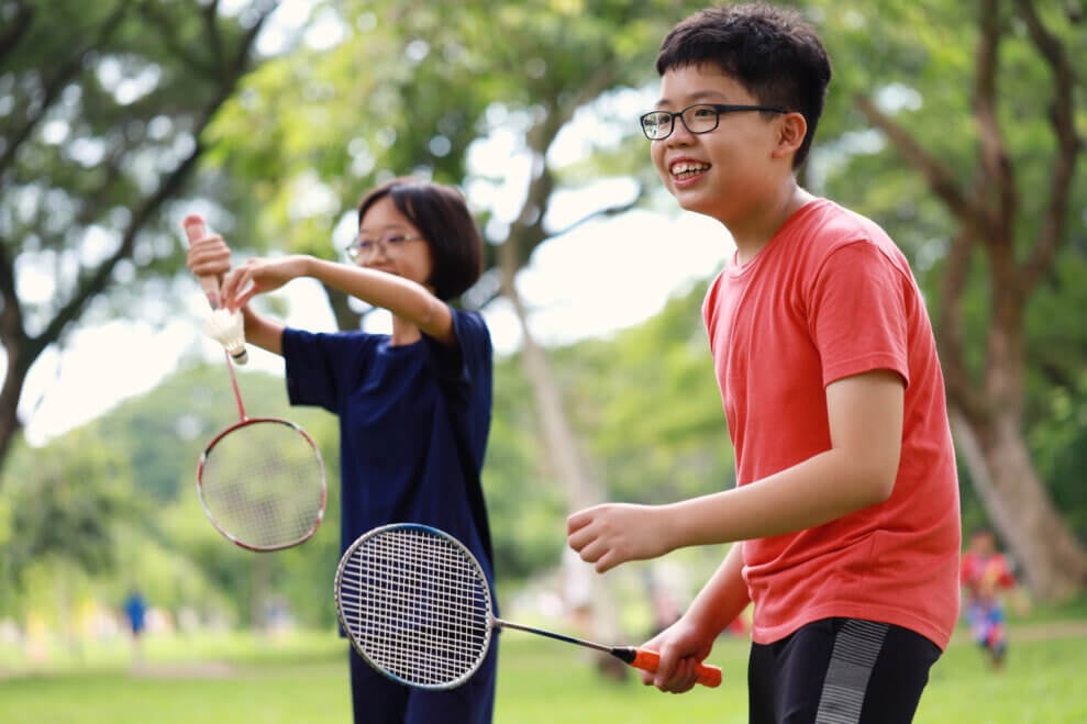 Two smiling children play badminton in a park.