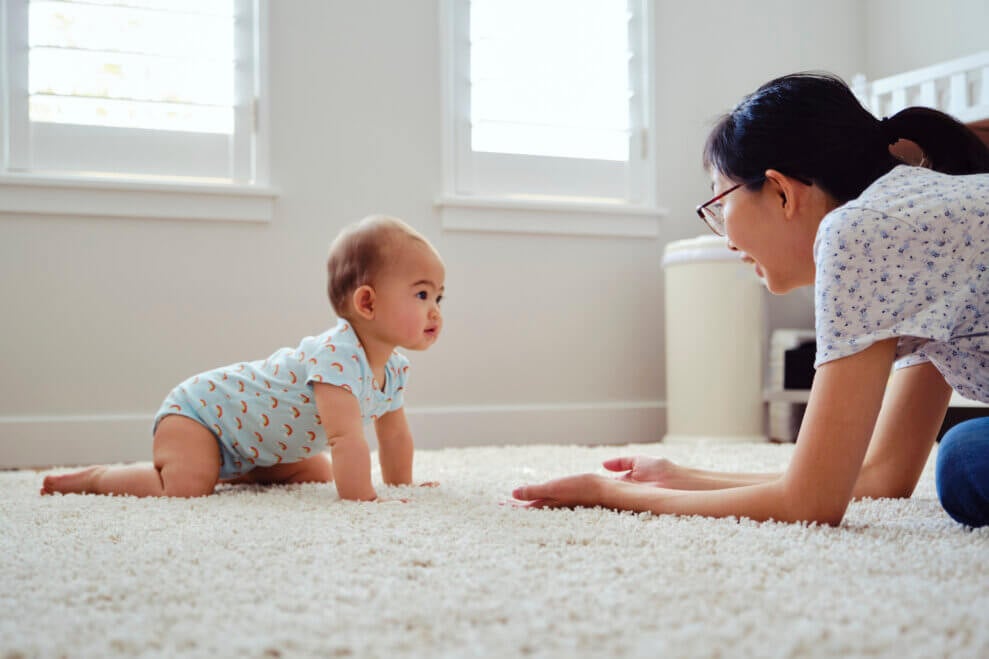 A mom sits in front of her baby on the carpet, encouraging the baby to crawl towards her.