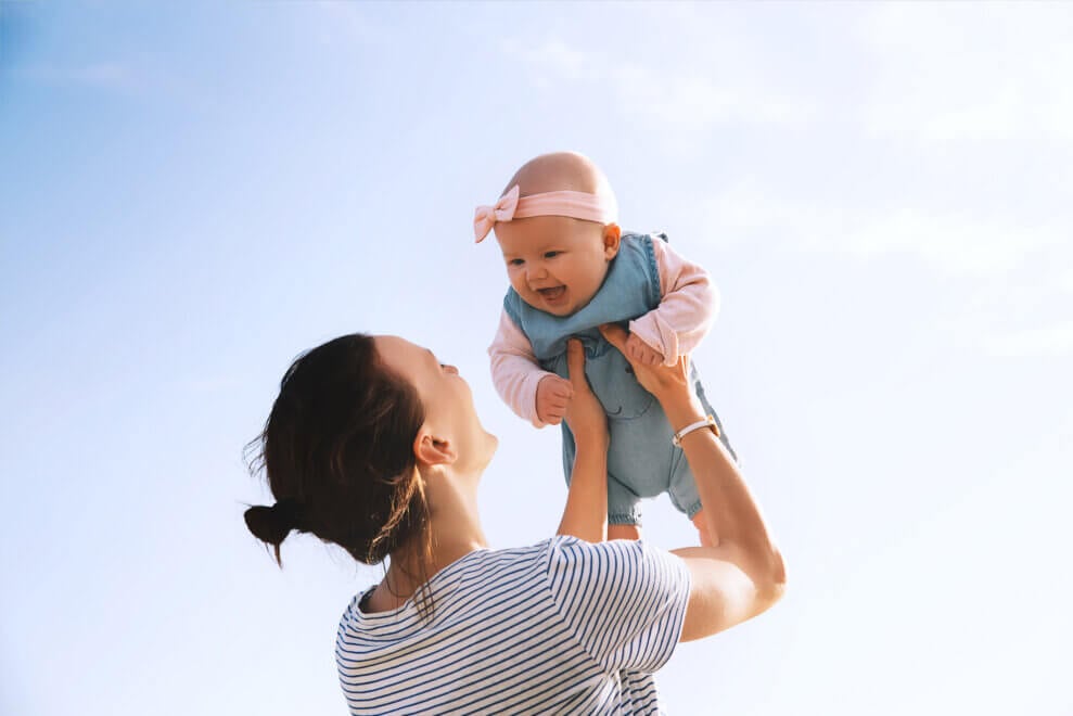 A mom holds her baby up in the air on a sunny day. Her daughter smiles down at her.