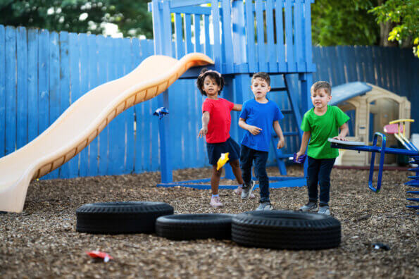 Three boys play bean bag toss outside on a playground.