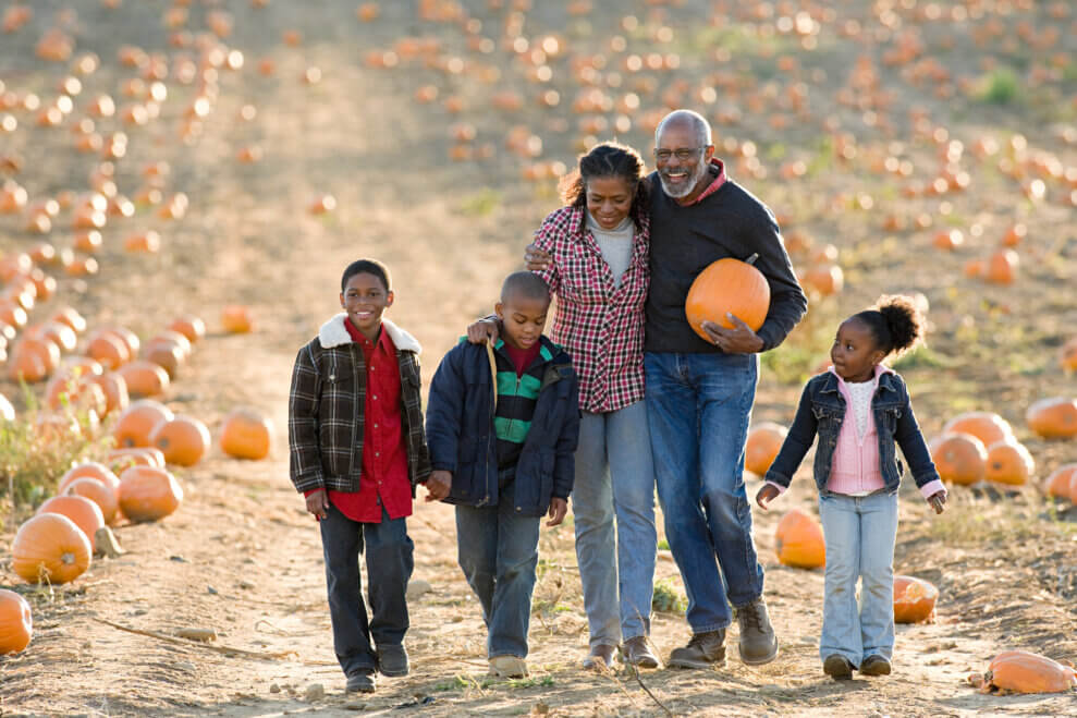 A father, mother, and their three kids walk together in the pumpkin patch on a fall day. The father carries a pumpkin in one arm and his other arm is around his partner's shoulders.