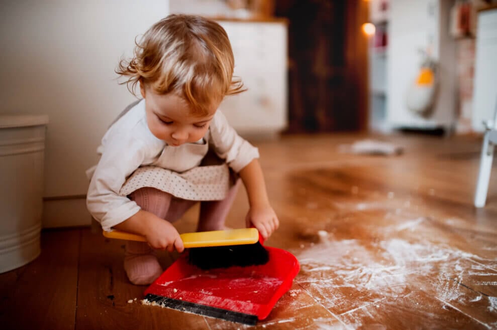 A toddler sweeps up flour on the floor using a small brush and dustpan.