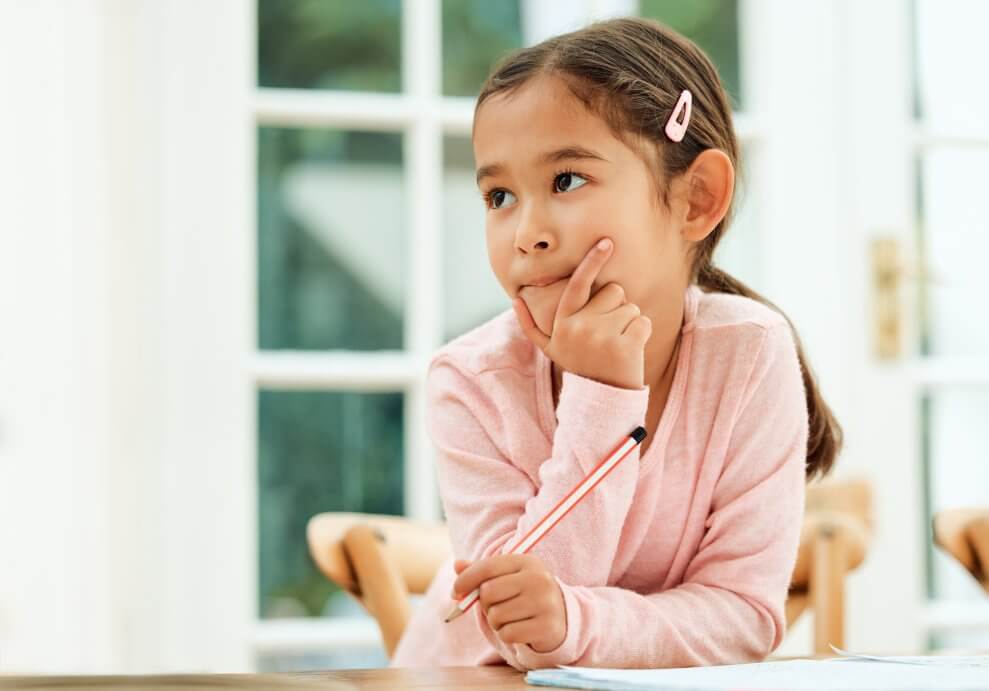 A girl sits with her journal at the kitchen table, with a pencil in her hand. She has her other hand on her chin and is deep in thought.