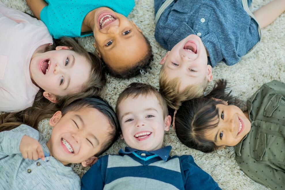 A group of young children lie on the floor with their heads forming a circle. They're smiling and laughing.