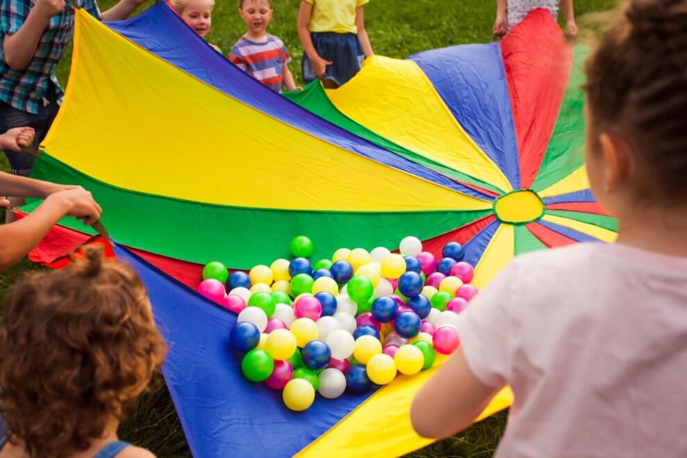 Many small multi-coloured balls rest in the middle of a parachute. A group of children holds the ends of the parachute up.