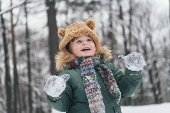 A young child wearing a toque, mitts, and a winter jacket laughs while standing outside in the snow.