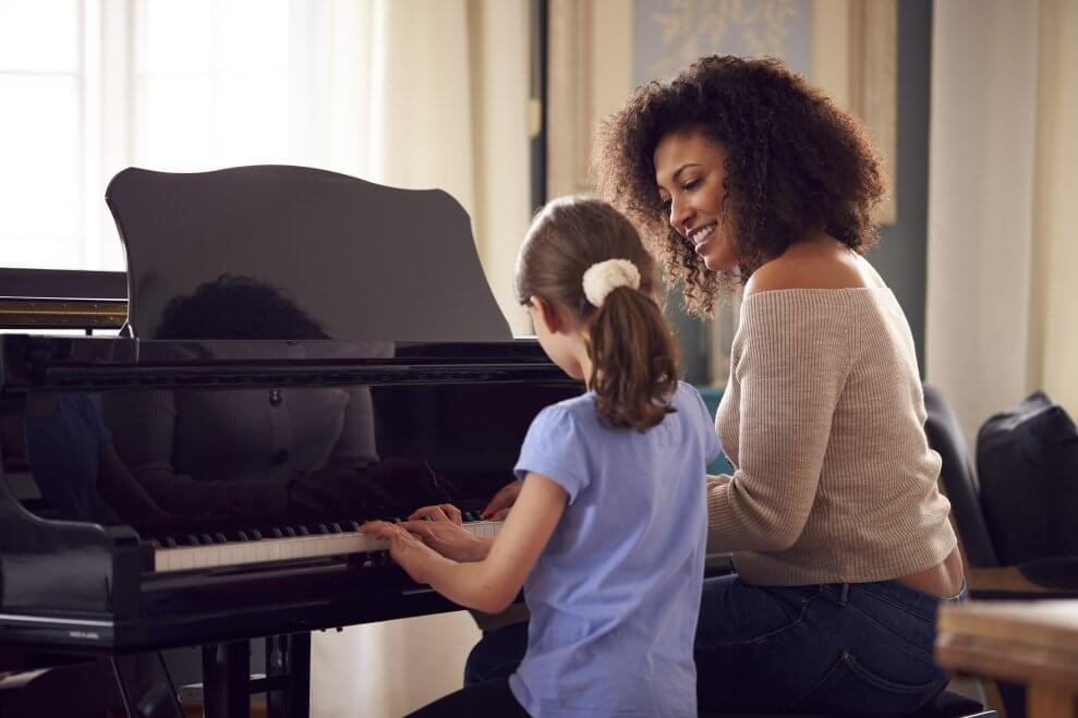 A girl and her piano teacher sit in front of a piano as the teacher shows her which keys to press.