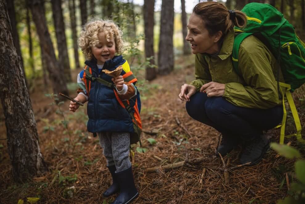 A young child holds up a mushroom to show her mother outside in the woods. The mother wears a backpack.