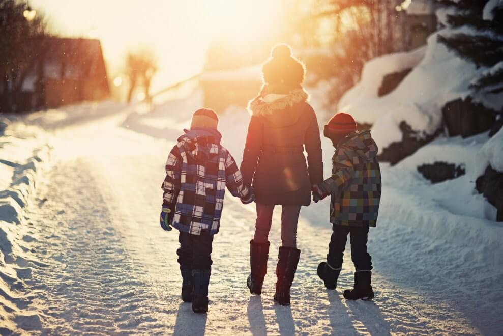 A mom walks hand-in-hand with her two children on a snowy path in the winter. The sun shines brightly on them.
