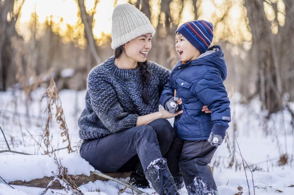 A mother sits on a log outside in the snow next to her young son, who's smiling. 