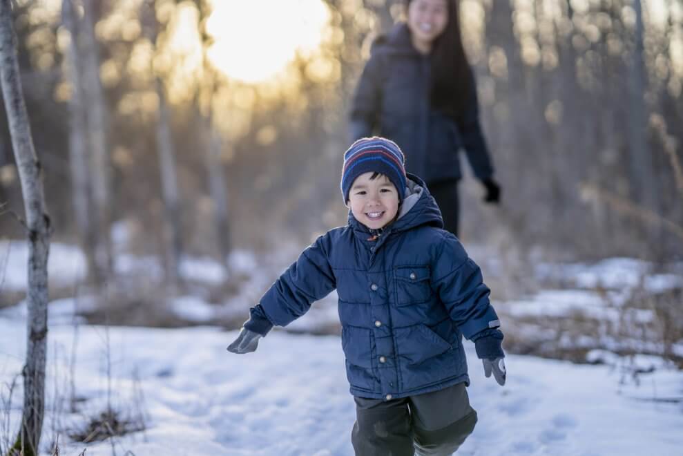 A young boy runs through the snow as his mom walks behind him, smiling.