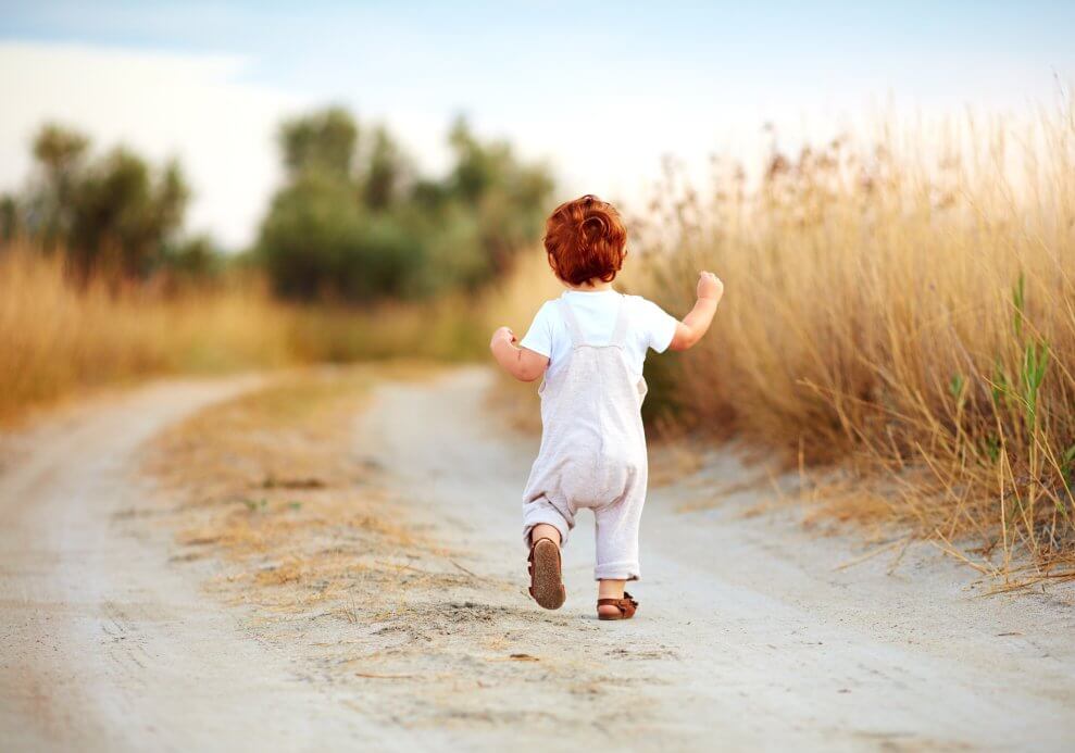A toddler wearing overalls and sandals runs along a path through a field.