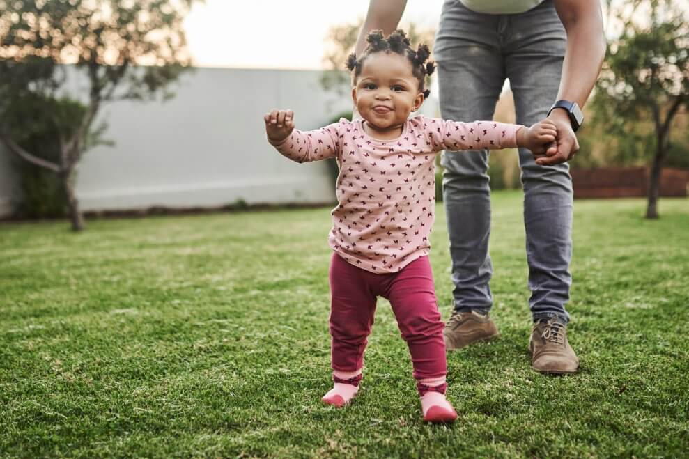 A toddler walks in her backyard, with a parent behind her holding her hand.