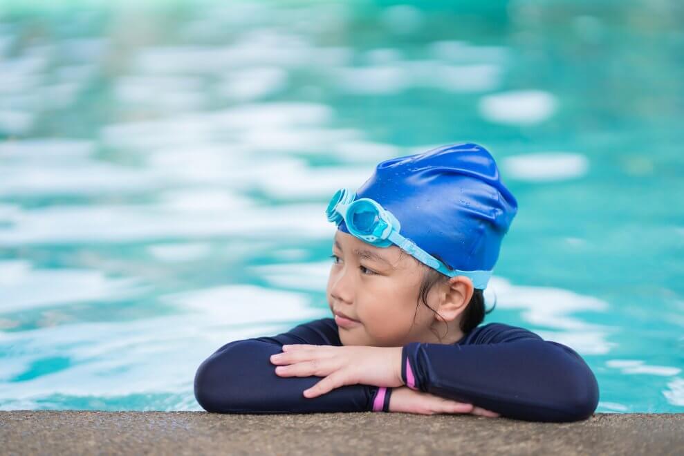 A young girl wearing a swim cap and goggles on top of her head rests her arms on the edge of a pool. 