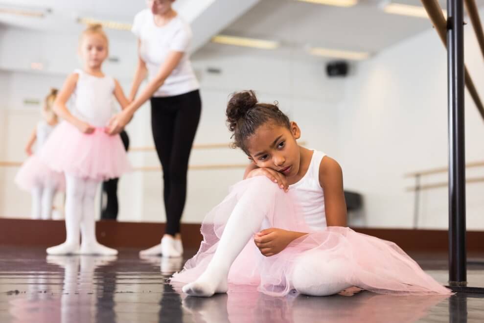 A girl sits on the floor of her ballet class, looking sad.