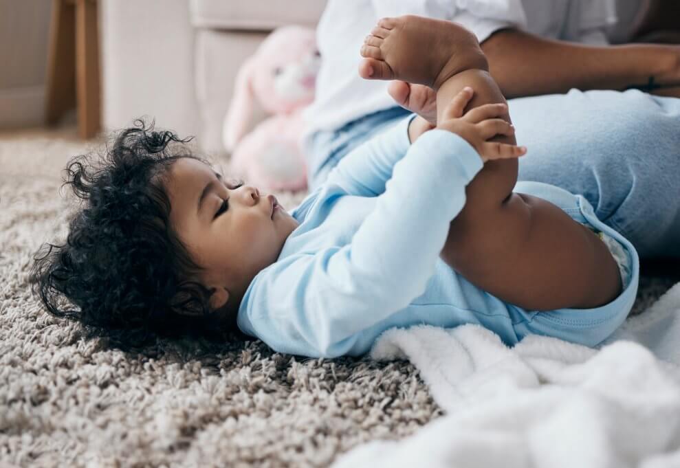 A baby lying on her back on the carpet touches her toes.