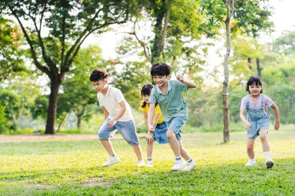 A group of four smiling kids runs outside in a grassy park on a sunny day.