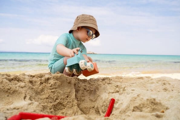A toddler boy plays on the beach wearing a sun hat and sunglasses.