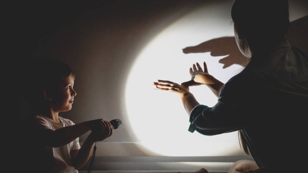 A mom and her son make shadow puppets on the wall in the dark using a flashlight.