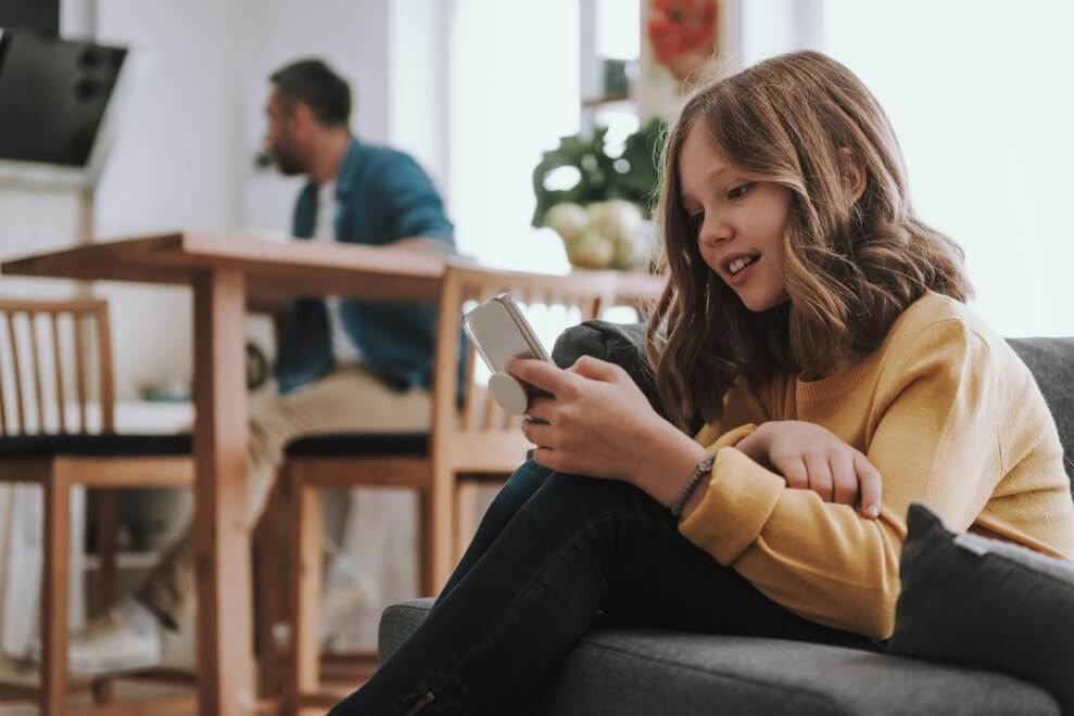 A preteen girls sits on the couch at home and scrolls on a smartphone.