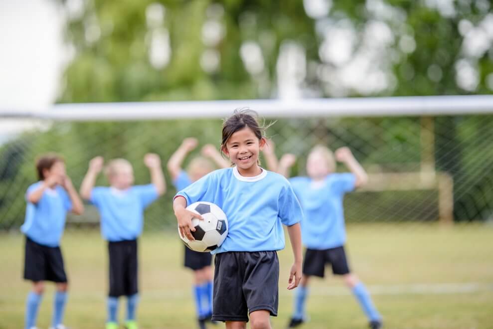A girl walks across the soccer field, holding a soccer ball, as her team cheers behind her.