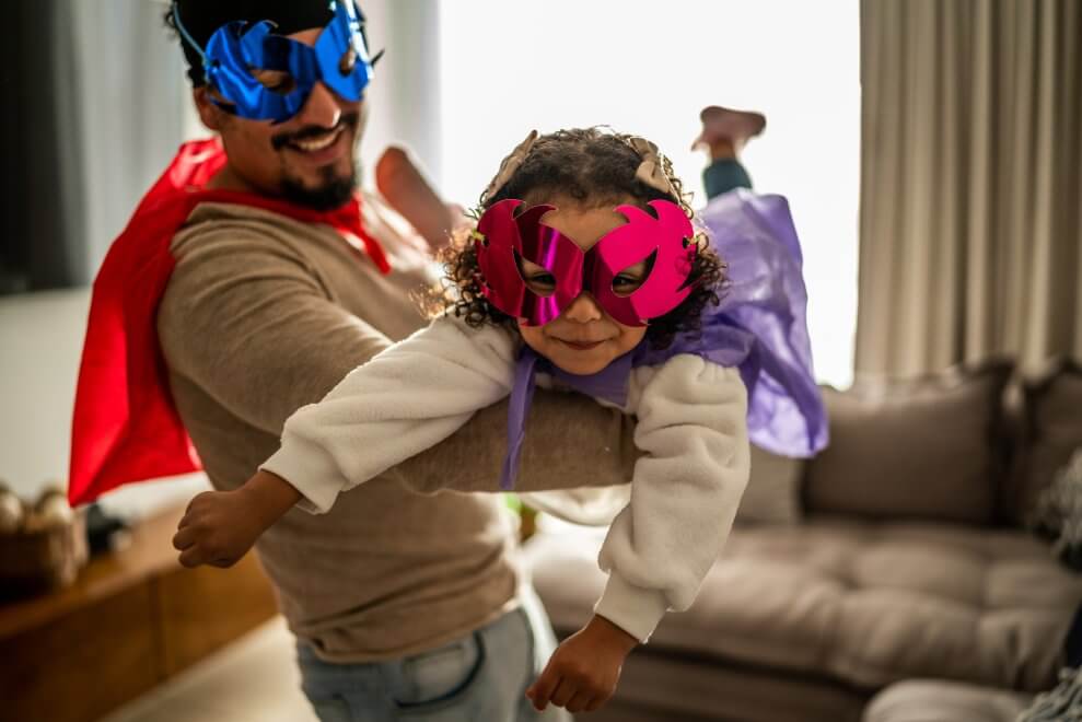 A father and his toddler daughter play airplane in the living room while dressed in masks and capes.