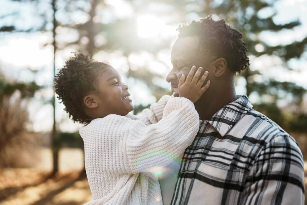 A father holds his toddler daughter in his arms outside while she pats his cheeks and smiles at him.