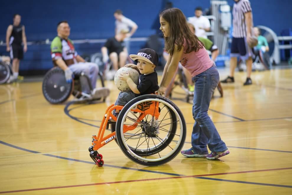 A girl pushes a young boy in an orange wheelchair around a gymnasium. He carries a ball on his lap.