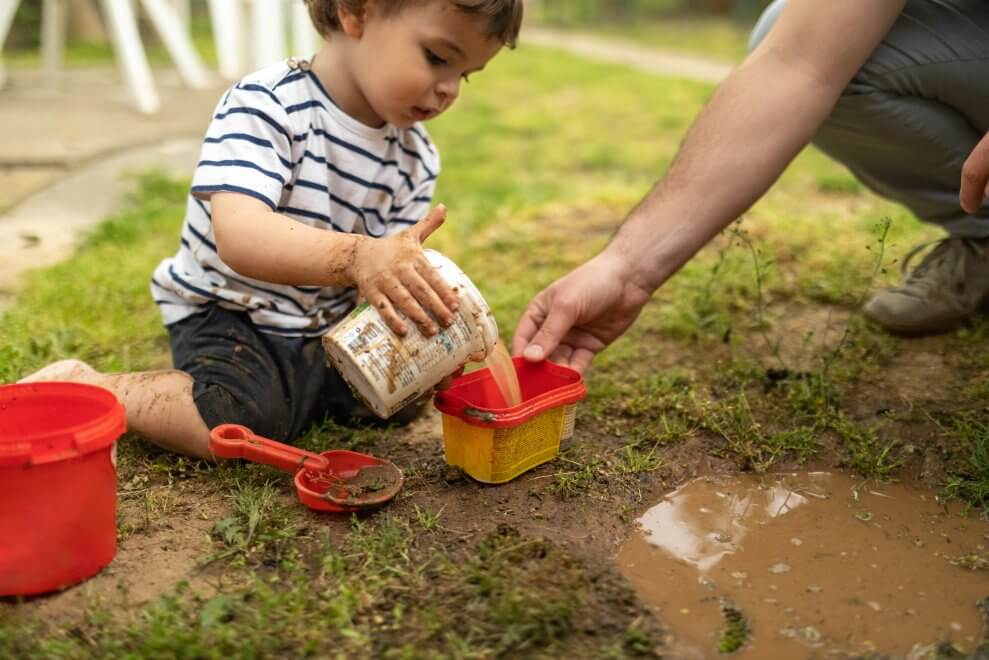 A toddler measures and pours dirt and water outside, helped by his father. Playing with mud and dirt can be a great STEM activity for kids.