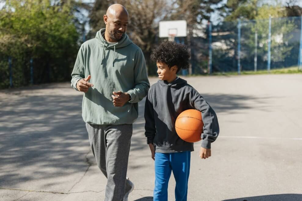 A dad and his teenage son walk across a basketball court, discussing a play. The son carries a basketball.